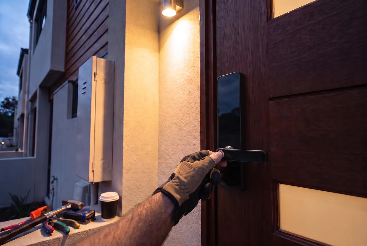 Gloved hand on the lever of a black keypad smart lock on a townhouse front door at dusk, with tools on the ledge